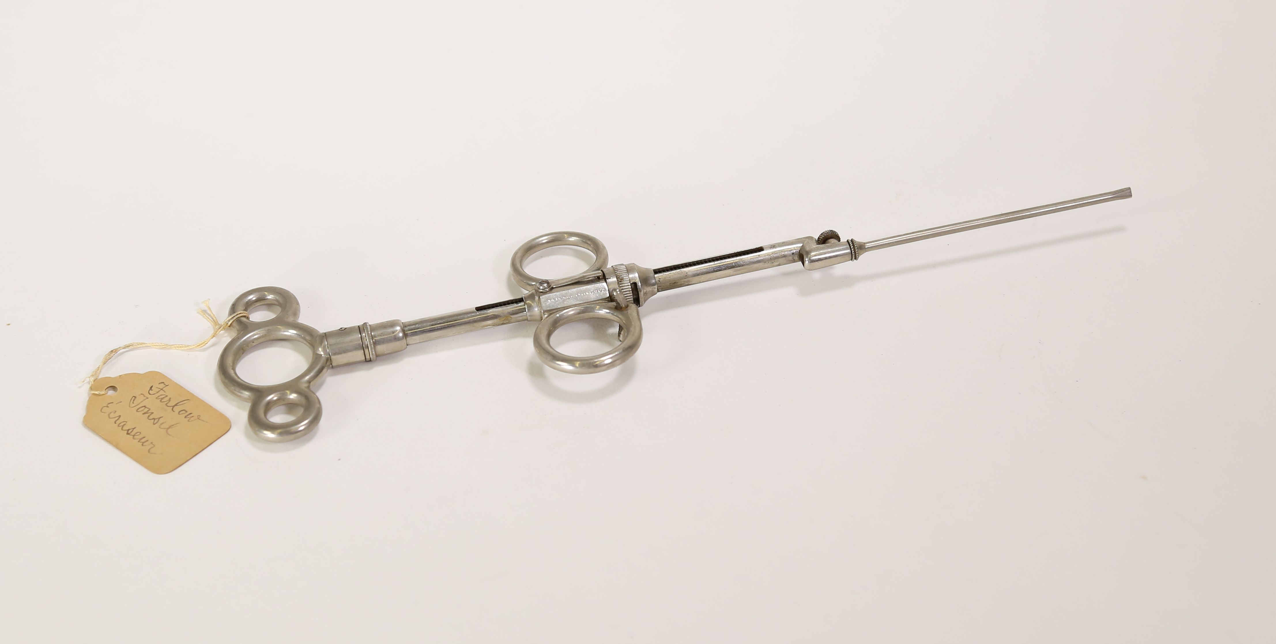 A slender, handheld metal surgical instrument with a label tied to the top that says Farlow Tonsil Écraseur.