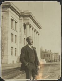 Levine standing in a formal pose in front of Gordon Hall, dressed in a three piece suit. His right hand is in his pocket; his left holds what appears to be a stethoscope.
