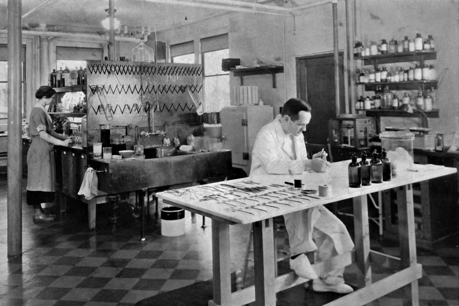 George van Siclen Smith sitting at a lab bench with instruments laid out beside him. Olive van Siclen Smith is standing at a lab bench in the background.