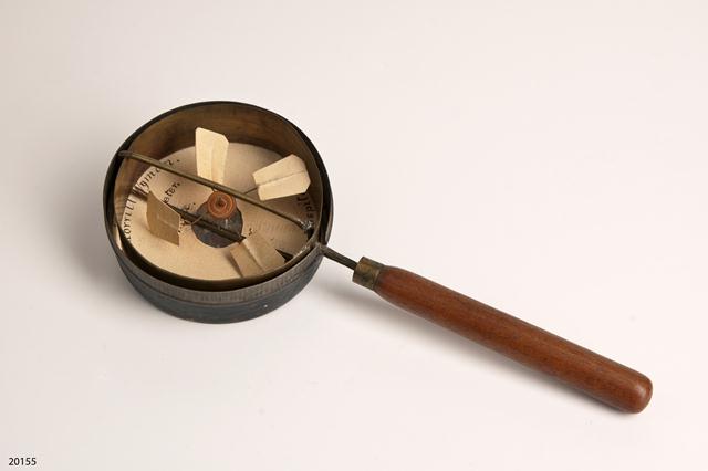 Wooden handled brass "airmeter" (anemometer). Airmeter is in metal case around fan section. Airmeter has open metal circle affixed to wooden handle. Metal circle has two diameter-long metal poles with small metal and paper fan (four pieces) inside. Round piece of paper sits in case describing airmeter.