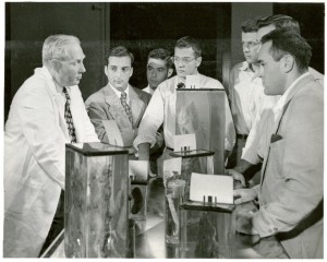 Dr. Hertig speaking to a group of 6 six students standing around several large wet specimens in rectangular glass containers.