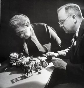 Black and white image of two people arranging miniature furnishings and figures