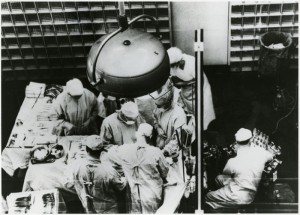 An operating room seen from above. Medical personnel attend to a patient next to tables covered with medical equipment.