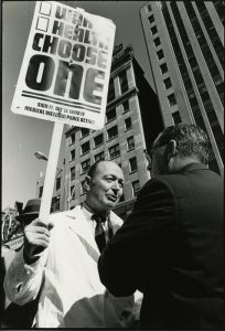Black and white image of HMS Dean Robert Ebert at a protest. Ebert is holding a sign that says "War Health Choose One" with check boxes next to War and Health. Ebert is talking to another man while holding the sign high.