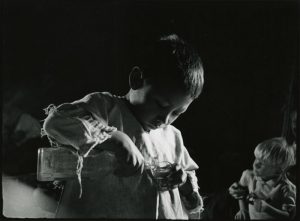 Black and white image of a Mayan child from Zinacantan wearing ragged clothes and pouring a drink from a bottle into a glass. There is another child is in the background.