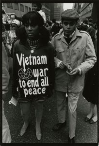  Black and white image of a woman and man protesting against the Vietnam War among a crowd of protestors. The woman is holding a sign that says "Vietnam War to end all peace."