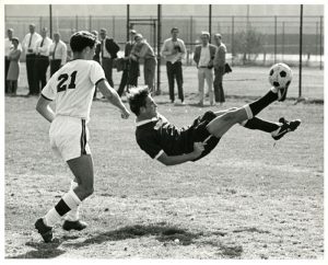Black and white action shot of a soccer match. One player is in a white uniform numbered 21 and is running towards another player in a black uniform. The player in the black uniform is hovering in the air and kicking a soccer ball.