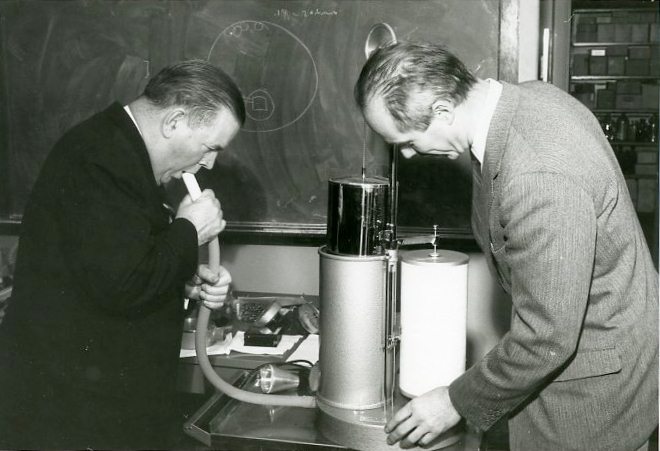 Two men demonstrating how to use a machine that measures lung function by breathing through a tube.