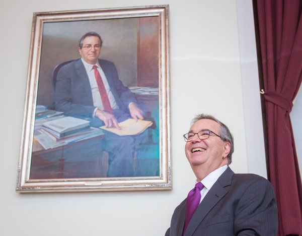 Jeffrey Flier smiles with a portrait of himself. In the portratit he sits at a desk in a suit with a book open across his lap.