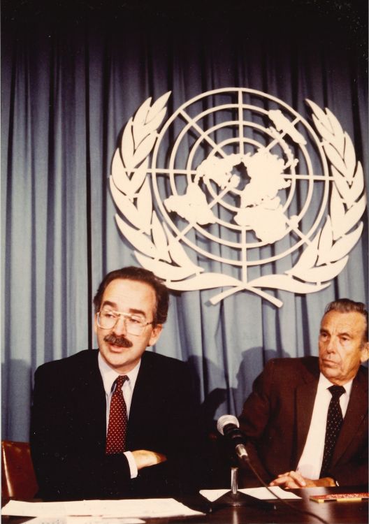 Jonathan Mann and another man sit at a table with a microphone in front of the UN logo.