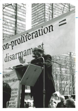 Man in a suit lectures at an outdoor podium in front of sign that says non-proliferation = disarmament