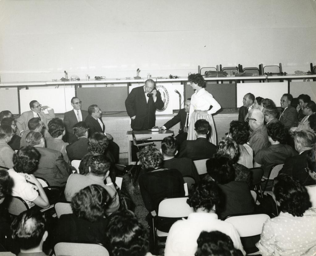 Black and white photograph of Jacob Moreno and Zerka Moreno standing next to a small desk in front of a seated audience of about thirty-six men and women. Jacob appears to be speaking into a telephone.