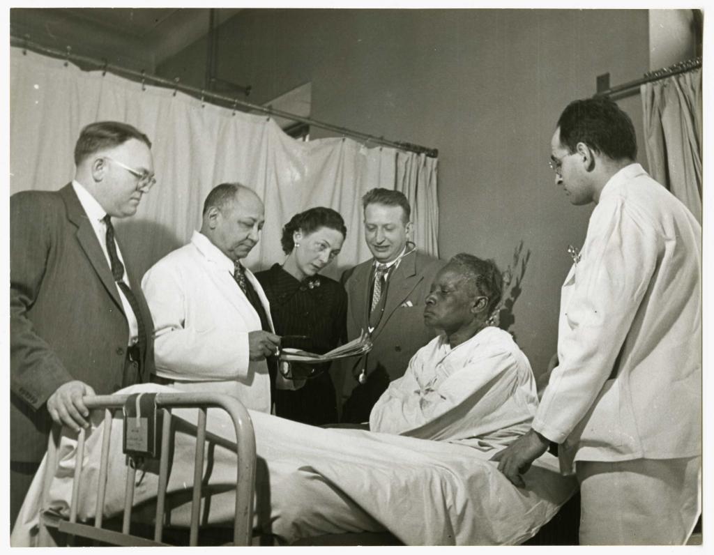 Black and white photograph of five medical professionals standing at the bedside of an African American woman, who is sitting up in the bed. Two of them are looking at a clipboard.