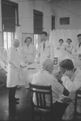 Doctors and nurses in white coats and uniforms gathered around a patient's bedside in a hospital room. In the foreground, two physicians are seated and appear to be conversing.