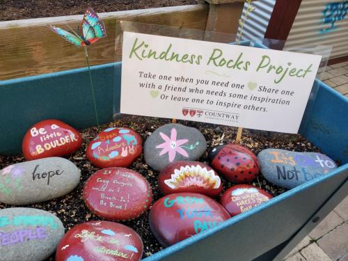 Photo of the Countway Kindness Rocks Garden (collection of colorful painted rocks) with a sign that reads "Kindness Rocks Project. Take one when you need one, share one with a friend who needs some inspiration, or leave one to inspire others."