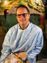 S. Abbas Shobeiri wearing a smock while holding an artist palette and paintbrush in his studio.