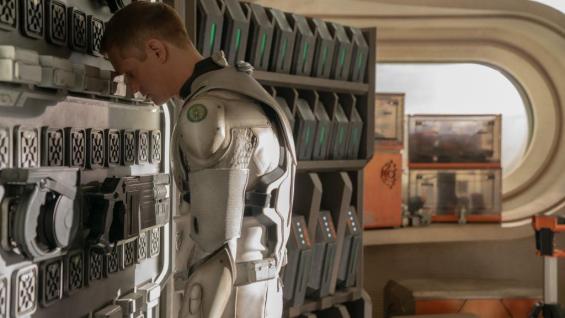 A cyborg standing in front a wall of futuristic technology slumps his shoulders and leans his human forehead against the wall with his eyes closed.
