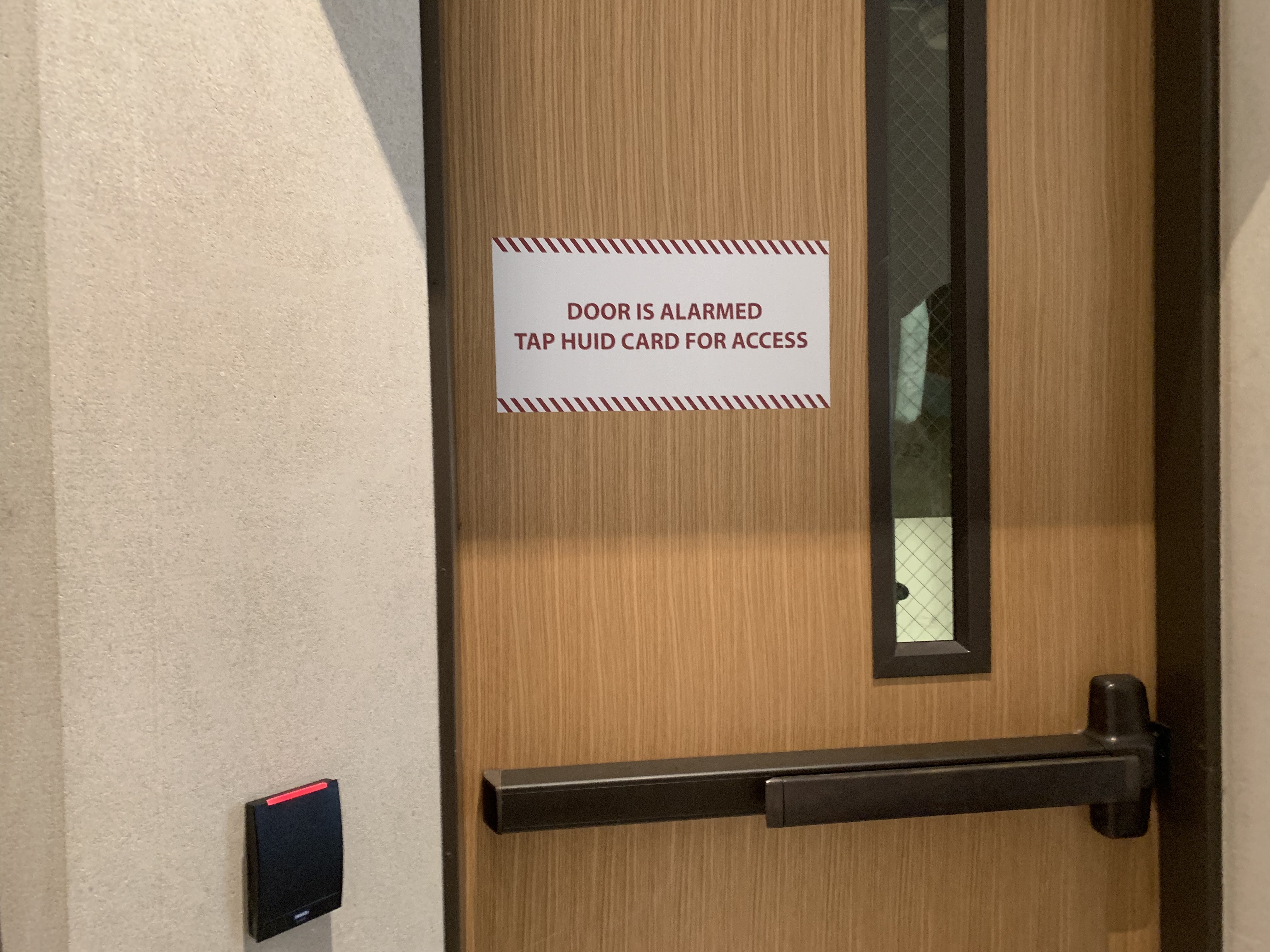 Wooden door next to a card reader with a sign that reads: Door is alarmed. Tap HUID card for more access.