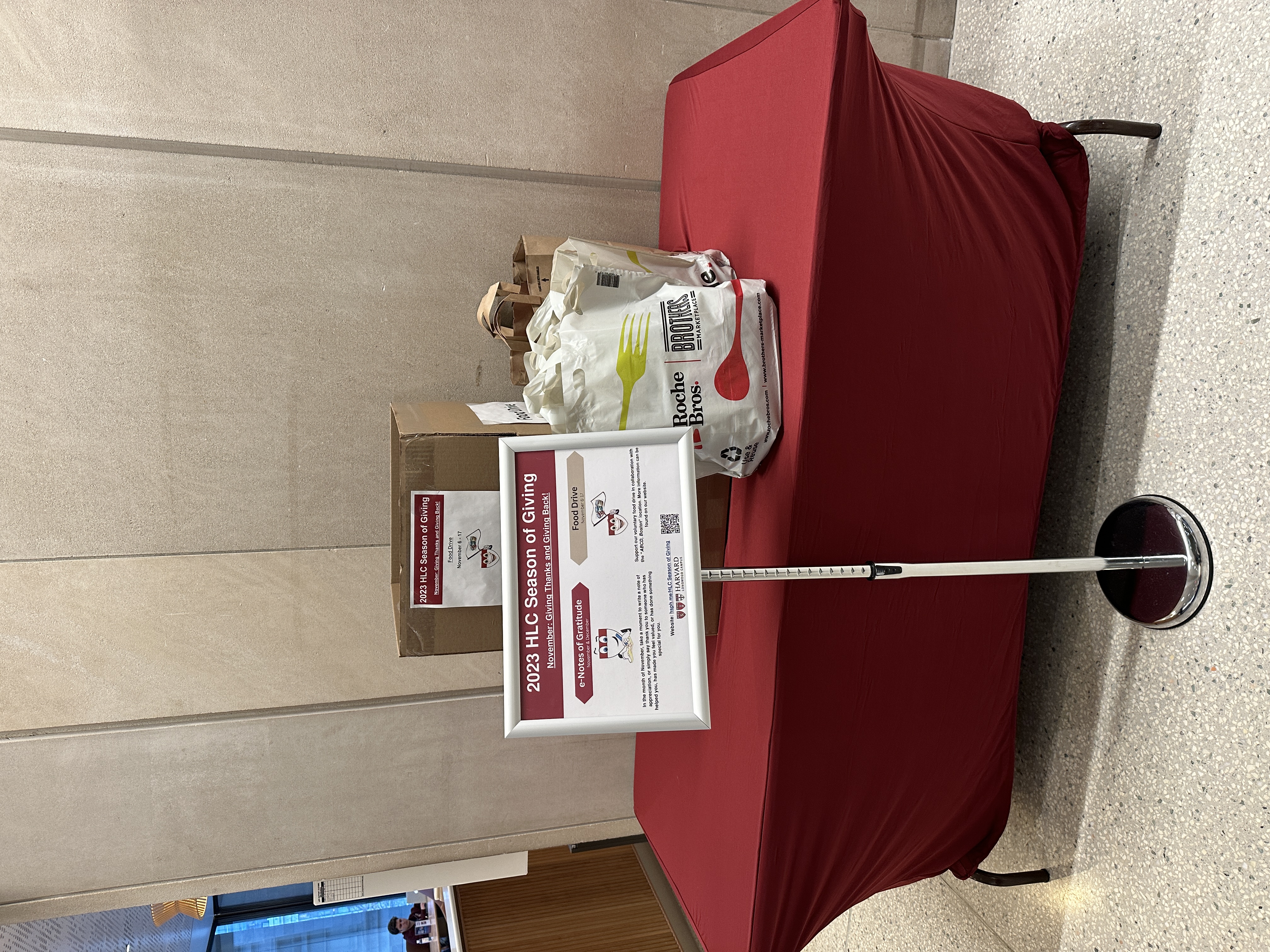 A table with boxes and bags behind the a sign that says: 2023 HLC Season of Giving; November: Giving Thanks and Giving Back. With details for two options: e-Notes of Gratitude and Food Drive.