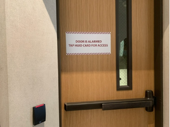 Sign on wooden stairwell door next to a card reader that says: Door is alarmed; Tap HUID card for access