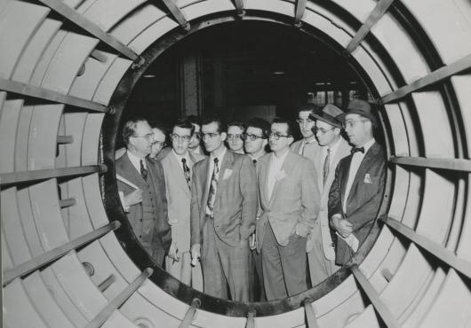 A group of white male students in suits and protective eyewear examine the inside of a large cylinder-shaped generator