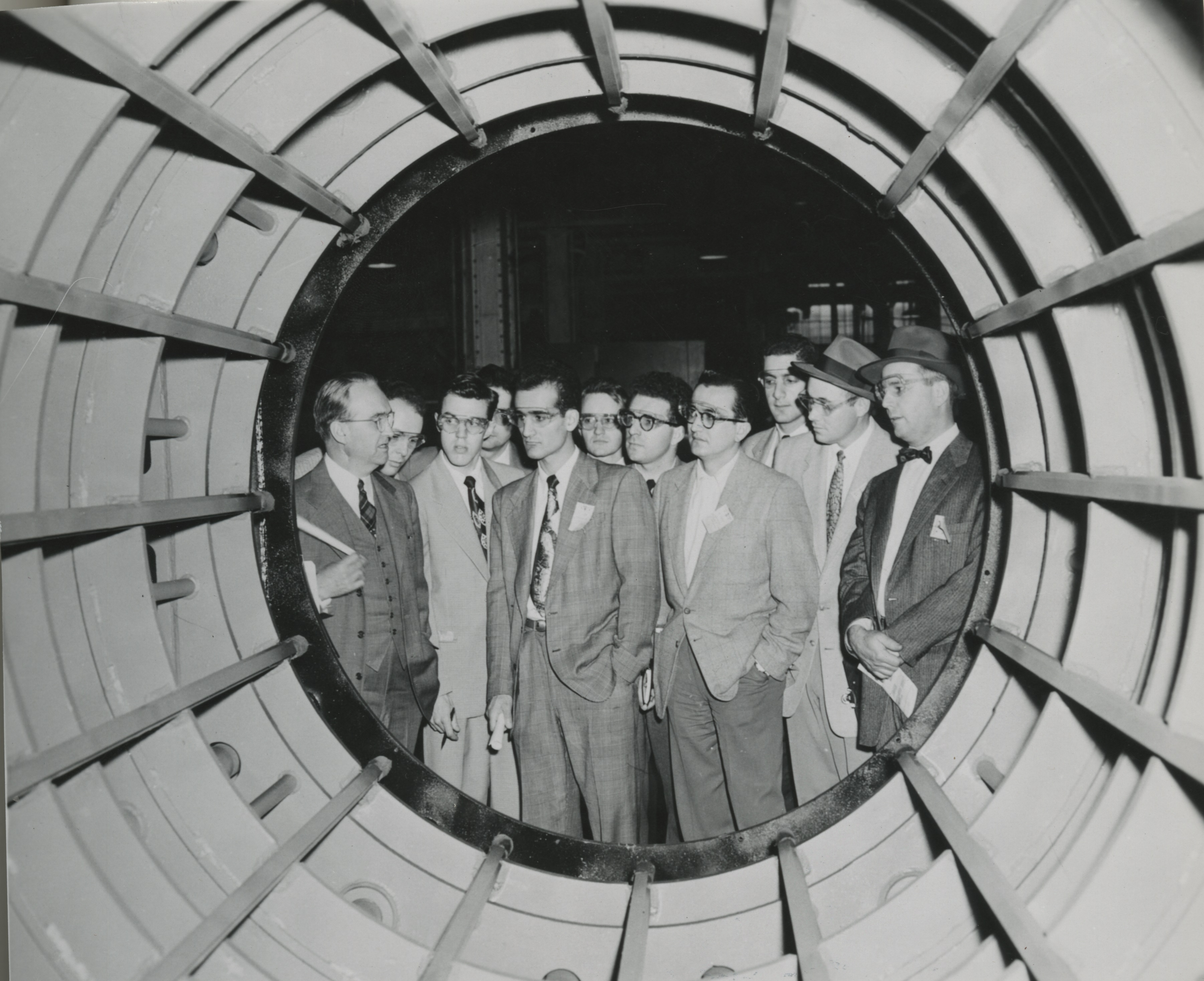A group of white male students in suits and protective eyewear examine the inside of a large cylinder-shaped generator