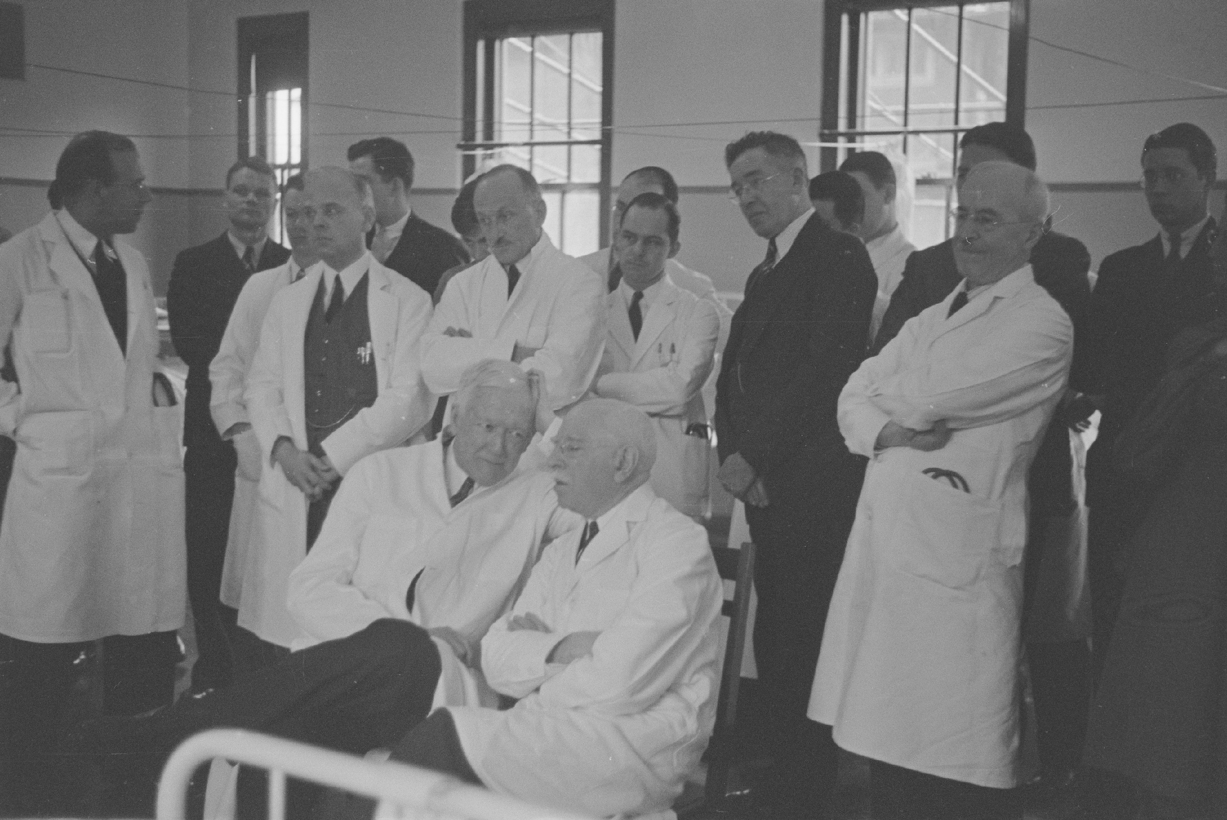 Group of doctors in suits or white coats sitting and standing around the end of a patient's bed in a hospital ward.
